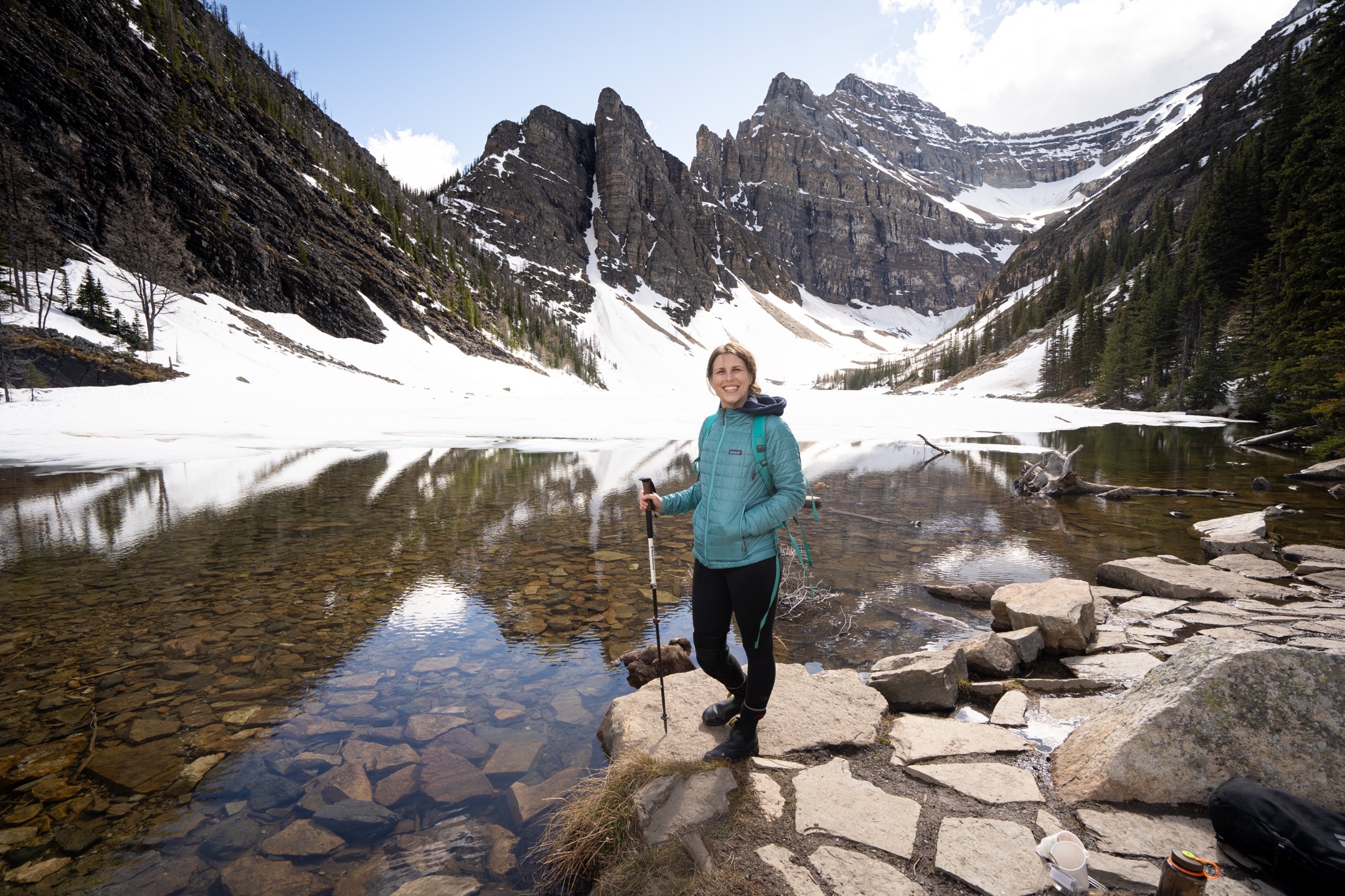 Standing on a rock at the edge of a clear alpine lake, holding a hiking pole, with snow-covered slopes and a jagged mountain ridge behind.